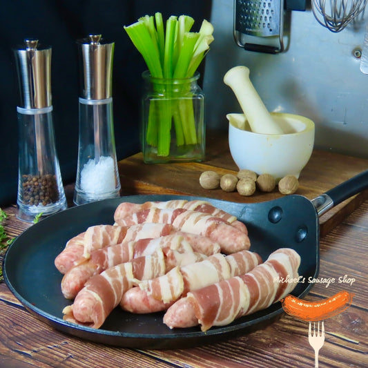 Bacon-wrapped sausages (pigs-in-blankets) on a pan with kitchen items in the background by Michael's Sausage Shop.