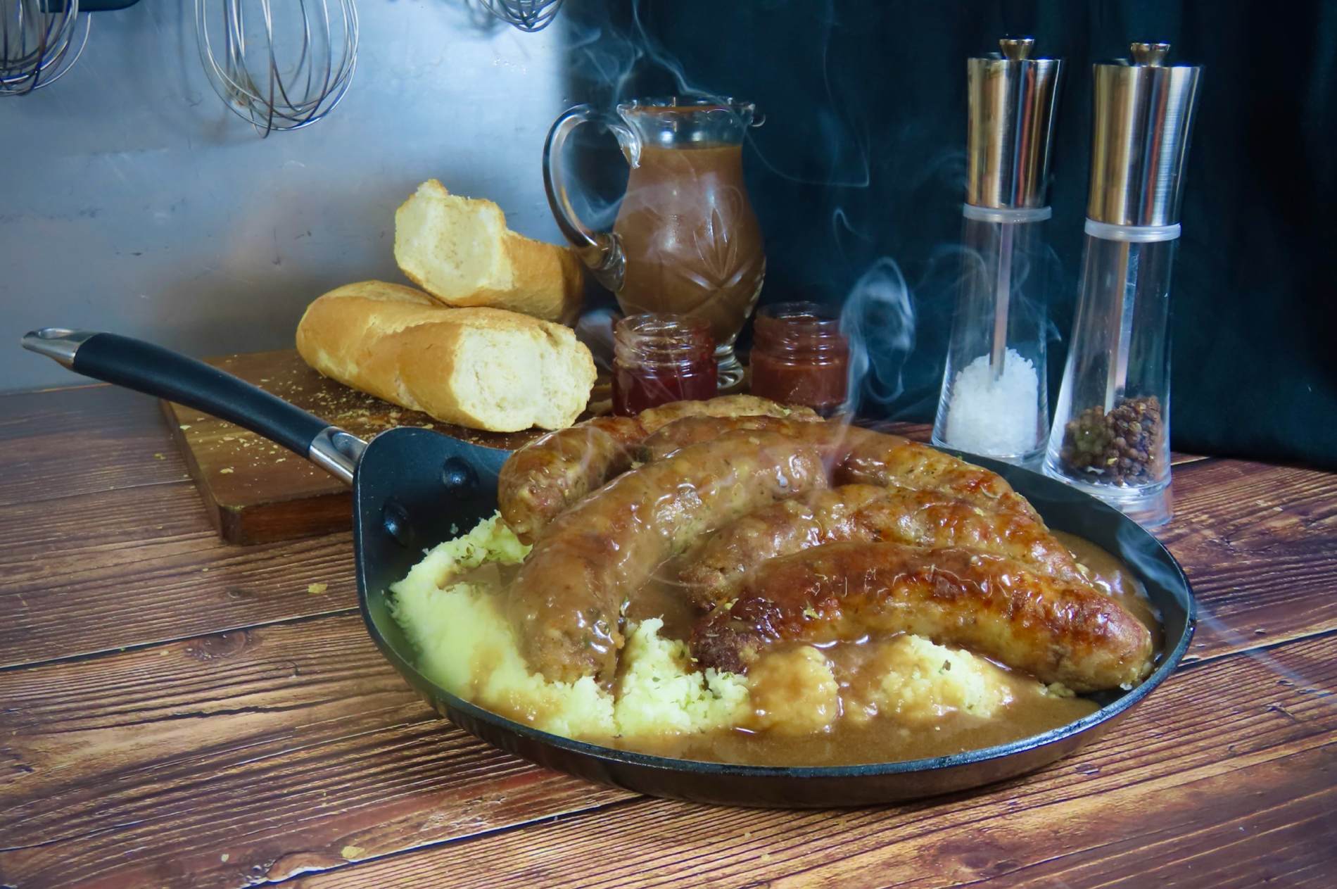 Sausage and mash dish in a pan on a wooden table with bread and condiments in the background. 